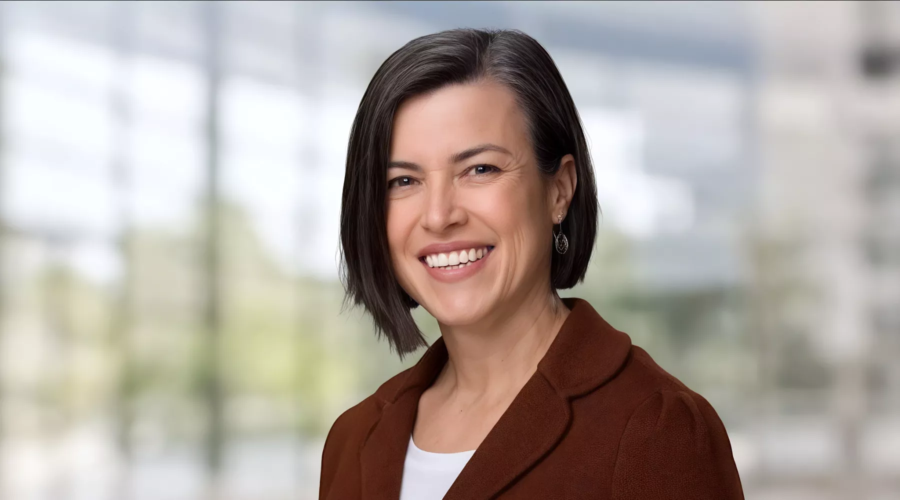 Dean Dayna Touron with short dark brown hair wearing a brown jacket and white top, smiling at the camera.