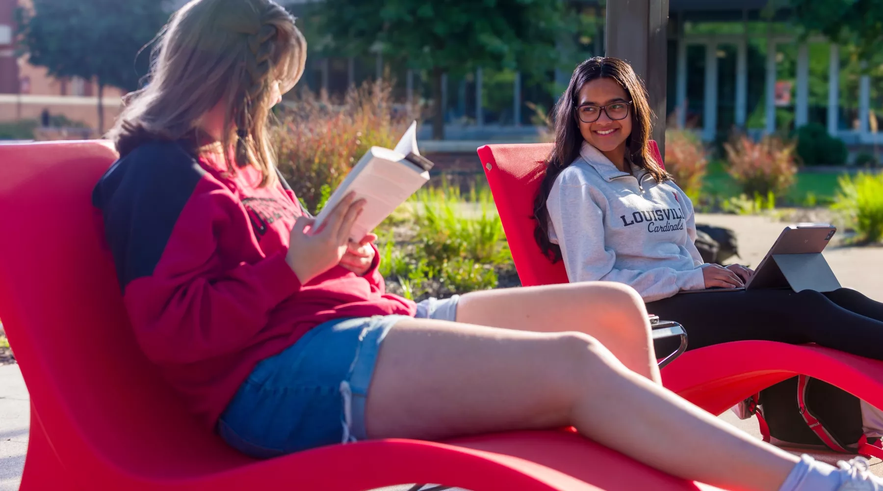 Two students outdoors on a sunny day takling while lounging in chairs