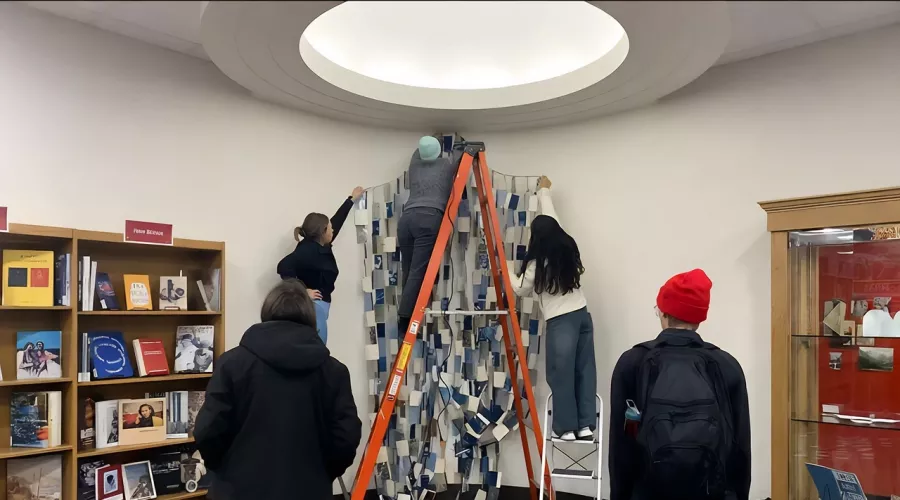 A group of individuals collaborates on a large fabric project inside a library setting, surrounded by books and resources.