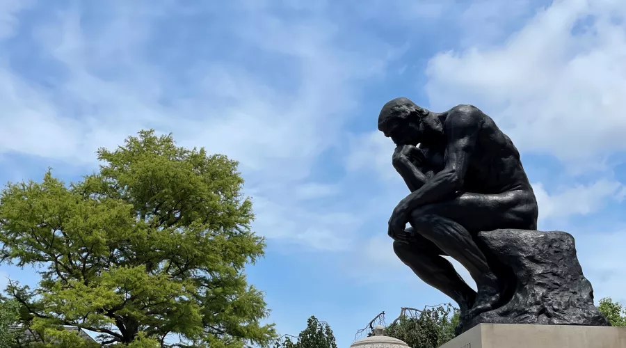UofL's thinker statue from the side with the sky in the background. Trees on campus can be seen in the background as well as a lamp post with a UofL banner.